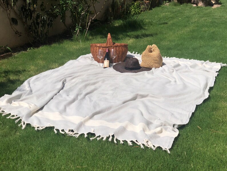 May include: A white picnic blanket with a gray stripe and fringe trim. A wicker picnic basket, a bottle of wine, two wine glasses, and two straw hats are on the blanket.