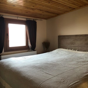 May include: A white and gray patterned bedspread on a bed with a wooden headboard and a wooden ceiling. The bed is in a bedroom with a window and brown curtains.