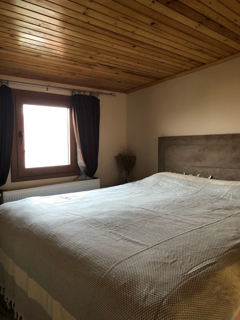 May include: A white and gray patterned bedspread on a bed with a wooden headboard and a wooden ceiling. The bed is in a bedroom with a window and brown curtains.