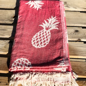 Puede incluir: Una toalla de playa roja con un patrón de piña blanca. La toalla tiene un borde de flecos.