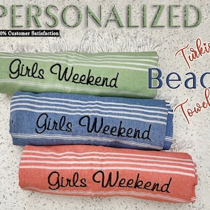 May include: Three Turkish beach towels rolled up, each with the text "Girls Weekend" embroidered in black. The towels are green, blue, and coral.