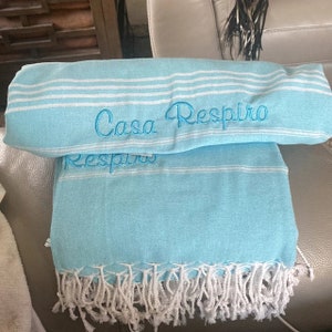 May include: A light blue beach towel with white stripes and fringe. The towel has the text "Casa Respiro" embroidered on it.