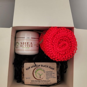 May include: A gift box set containing raw African black soap, shea butter, and a red washcloth. The soap bar is wrapped in brown paper with the text "Raw African Black Soap" and "Organic Shea". The shea butter is in a white jar with the text "Shea Butter".