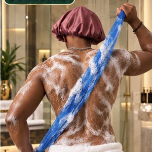 May include: A long, blue, textured exfoliating back scrubber strap measuring 50 inches in length. The strap is covered in white soap suds and is being used to scrub a back. The individual is wearing a mauve satin shower cap and a white towel.