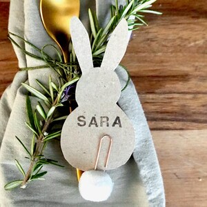 Könnte beinhalten: Eine österliche Tischdekoration mit einer hasenförmigen Tischkarte mit dem Namen "SARA". Die Tischkarte ist mit einem goldenen Löffel und Rosmarinzweigen an einer Serviette befestigt. Der Hase hat einen weißen Baumwollschwanz.