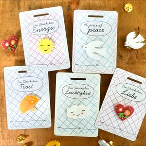 Könnte beinhalten: Fünf dekorative Anstecknadeln auf einzelnen Präsentationskarten. Die Anstecker zeigen eine gelbe Sonne, eine weiße Taube, einen orangefarbenen Regenschirm, eine weiße Wolke und ein rotes Herz. Jede Karte hat ein Gittermuster und die Aufschriften Ein Stückchen Energie, A piece of peace, Ein Stückchen Trost, Ein Stückchen Leichtigkeit und Ein Stückchen Liebe.
