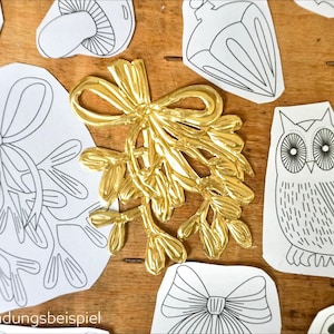 May include: A shiny gold-colored metallic mistletoe ornament with a bow, surrounded by various black and white paper cutouts featuring line drawings of an owl, a mushroom, a bow, and other botanical shapes on a wooden surface.