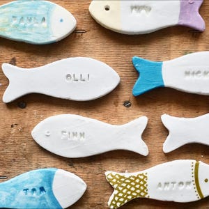 Op de afbeelding: Handgeschilderde witte keramische vissen met namen als "Tom" en "Finn". Elke vis heeft een uniek kleurenschema, met blauwe, groene en gele accenten. De vissen zijn geplaatst op een houten oppervlak.