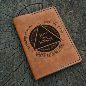 Puede incluir: Un portapasaporte de cuero marrón con un diseño grabado. El diseño presenta un triángulo con el texto "Party Like Hobbits" en su interior. El triángulo está rodeado por un círculo con el texto "Drink Like Dwarves, Smoke Like Men, Sing Like Elves".