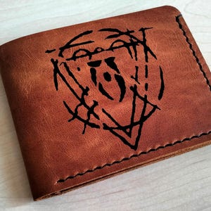 May include: A brown leather wallet featuring a black graphic design. The wallet is rectangular with a stitched border. The leather has a natural, textured appearance, typical of quality craftsmanship.