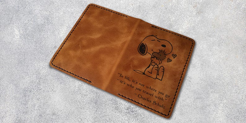 May include: A brown leather passport holder with a quote from Charles Schulz about travel and a cartoon image of Snoopy hugging Woodstock.