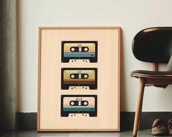 May include: A framed print featuring three retro cassette tapes in teal, yellow, and pink. The tapes are arranged vertically against a light beige background, encased in a wooden frame. The print is displayed on a tiled floor, near a black chair with a gray blanket.