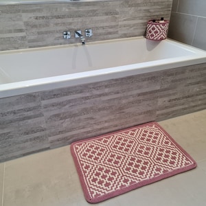 Può includere: Una vasca da bagno bianca con un tappetino da bagno rosa di fronte. La vasca da bagno è posizionata contro una parete piastrellata con uno schema di colori grigio e bianco.