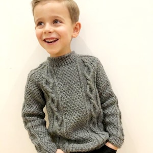 Pode incluir: Um suéter cinza de malha com um padrão de trança. O suéter tem um decote redondo e mangas compridas.