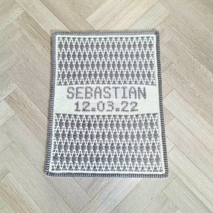Puede incluir: Una manta de bebé de crochet blanca y gris con el nombre "Sebastian" y la fecha "12.03.22" bordada en gris.