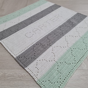 May include: A crocheted baby blanket with stripes of white, grey, and light green. The blanket has a simple, repeating pattern of hearts.