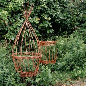 May include: Two brown woven willow plant supports. The taller support features a conical, teepee-like structure with vertical rods gathered at the top. The shorter support is cylindrical with a woven base and sides, both designed for garden use.