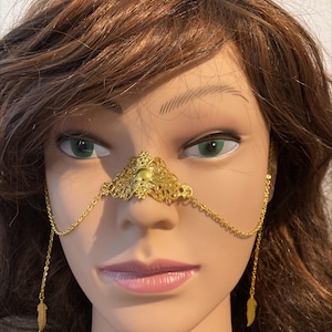 May include: Gold nose chain with a decorative floral design and feather accents. The chain is attached to a nose ring.