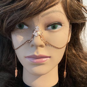 May include: A gold nose chain with a decorative design and feather charms. The chain is worn on a mannequin head.