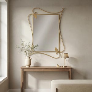 Pode incluir: Um espelho de parede retangular com uma moldura de metal dourado com volutas assimétricas fluidas e detalhes em forma de folha. O espelho está montado sobre uma mesa de console de madeira que contém um vaso de cerâmica com plantas e uma pequena lâmpada.