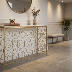 Puede incluir: Una mesa consola rectangular con una superficie de mármol blanco y una estructura de metal dorado con un intrincado diseño calado de espirales interconectadas. La mesa se apoya sobre cuatro patas delgadas y sostiene un libro marrón y un jarrón.