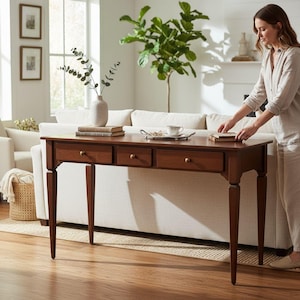 Può includere: Un tavolo console rettangolare in legno con tre cassetti dotati di piccoli pomelli rotondi in ottone, sostenuto da quattro gambe affusolate. Il piano del tavolo ospita un vaso in ceramica bianca con steli di eucalipto, libri e un piccolo vassoio argentato.