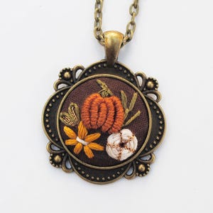 May include: A necklace featuring a round, bronze-toned metal pendant with an ornate, scalloped edge. The center contains dark brown fabric embroidered with an orange pumpkin, a white pumpkin, and a yellow flower with green stems.