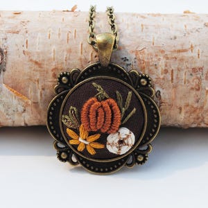 May include: A round pendant necklace featuring an antique bronze-toned metal frame with ornate detailing. The center displays dark brown fabric embroidered with a textured orange pumpkin, a small white flower, and a yellow flower with green stems.
