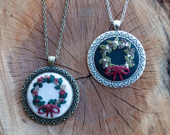 May include: Two silver and bronze necklaces with embroidered Christmas wreaths. One wreath is on a white background with red and green accents, and the other is on a black background with red and green accents.