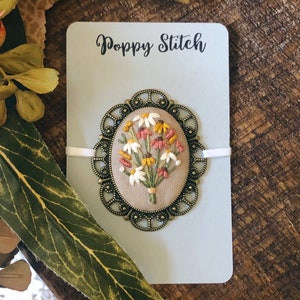May include: A hand-stitched floral brooch with a variety of white, yellow, and pink flowers in a bronze oval frame. The brooch is on a light blue card with the text "Poppy Stitch" printed in black.