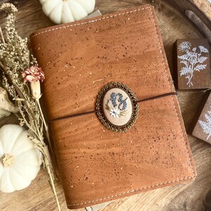 May include: A brown cork notebook with a stitched edge and a brown elastic band closure. A small, round, embroidered design with white flowers and a blue letter "B" is attached to the front cover.