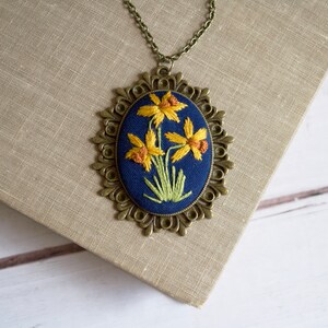 K&ouml;nnte beinhalten: Eine goldfarbene Halskette mit einem blauen ovalen Anh&auml;nger, der mit gelben Narzissen bestickt ist.