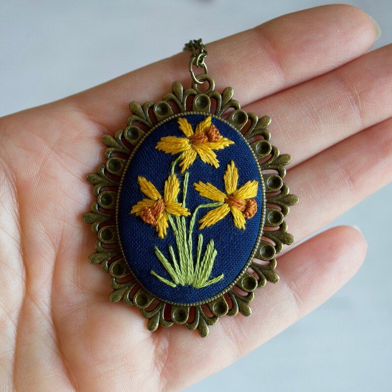 K&ouml;nnte beinhalten: Eine goldfarbene Halskette mit einem Anh&auml;nger aus blauem Stoff, der mit gelben Narzissen bestickt ist. Der Anh&auml;nger ist von einem dekorativen, verzierten Rahmen umgeben.