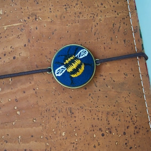 May include: A blue fabric circle with a yellow and black embroidered bee design. The circle is attached to a brown elastic band.