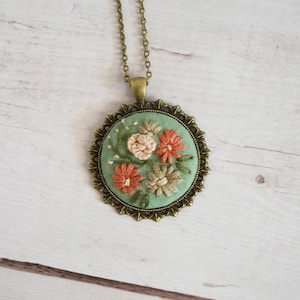 May include: A delicate floral necklace with a green embroidered pendant featuring pink and white flowers in a bronze oval frame.