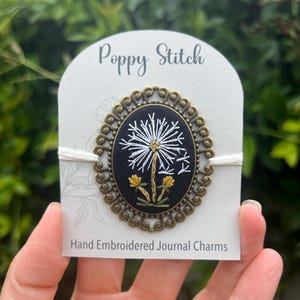 May include: An oval, hand-embroidered journal charm featuring a dandelion design. The charm has a black background with white and yellow thread details. The charm is set in a decorative bronze-colored frame and is attached to a white card that reads "Poppy Stitch" and "Hand Embroidered Journal Charms."