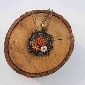 May include: A round pendant necklace featuring an embroidered design on dark brown fabric. The embroidery depicts an orange pumpkin, a white cotton boll, and a yellow flower. The pendant has an antique bronze-toned metal frame and chain.