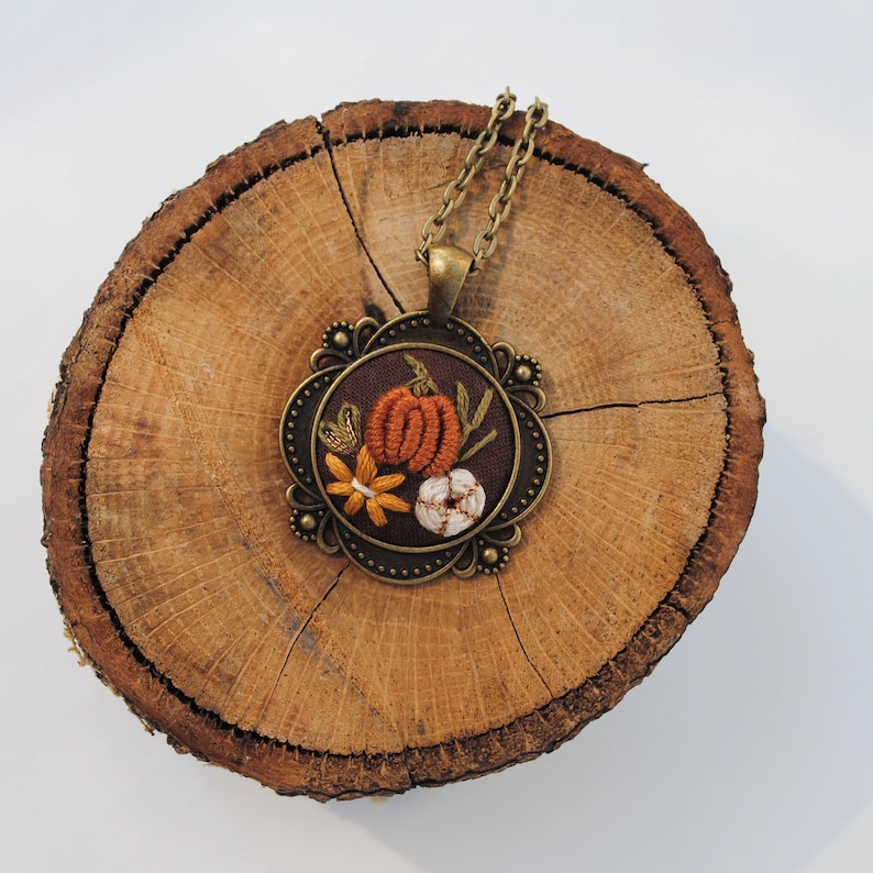 May include: A round pendant necklace featuring an embroidered design on dark brown fabric. The embroidery depicts an orange pumpkin, a white cotton boll, and a yellow flower. The pendant has an antique bronze-toned metal frame and chain.
