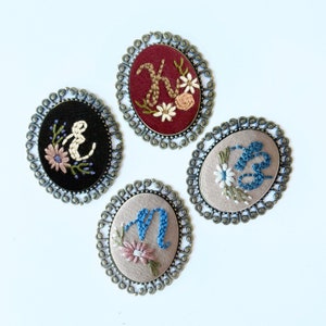 May include: Four embroidered brooches with different letters and floral designs. Each brooch is in a gold-toned oval frame with a decorative edge.