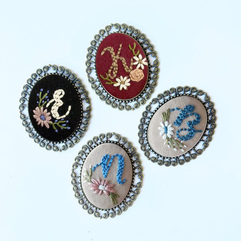 May include: Four embroidered brooches with different letters and floral designs. Each brooch is in a gold-toned oval frame with a decorative edge.