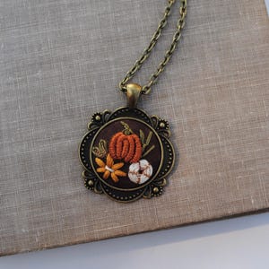 May include: A necklace featuring a round, dark brown fabric pendant with an embroidered orange pumpkin, a yellow flower, and a white cotton boll. The pendant is set in an ornate, antique bronze-toned metal frame and hangs from a matching chain.