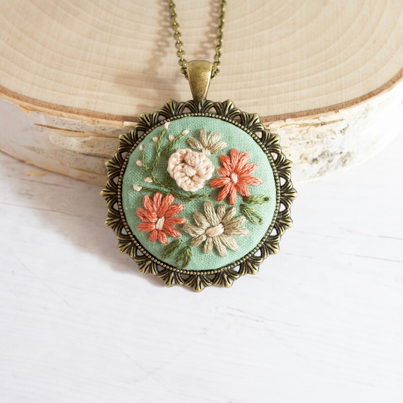May include: A green fabric pendant with embroidered flowers in shades of pink, peach, and white, framed in a bronze-colored metal setting. The pendant is attached to a bronze chain.