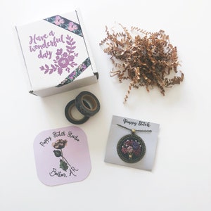 May include: A white cardboard box with floral tape and the text Have a wonderful day, brown paper filler, two rolls of dark patterned washi tape, a square card with a flower illustration and Poppy Stitch Studio Benton, AR text, and a necklace.
