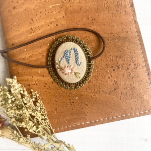 May include: A brown leather journal with a decorative metal bookmark featuring an embroidered letter "M" with pink and blue flowers.