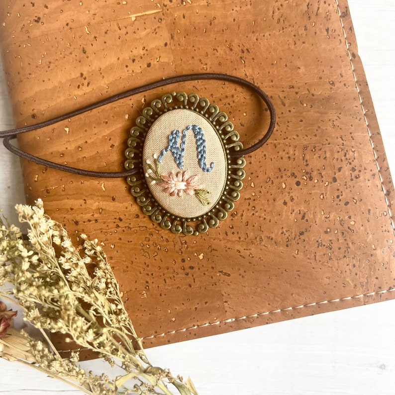 May include: A brown leather journal with a decorative metal bookmark featuring an embroidered letter "M" with pink and blue flowers.