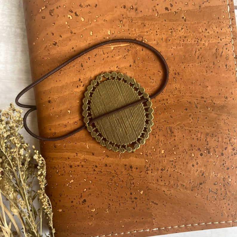 May include: A brown cork notebook with a gold-colored metal bookmark attached to it with a brown elastic band.