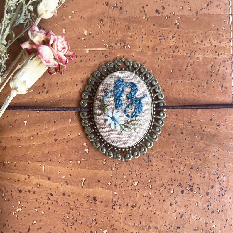May include: A small, round, embroidered pin with a blue and white floral design. The pin is surrounded by a decorative, gold-colored metal frame.