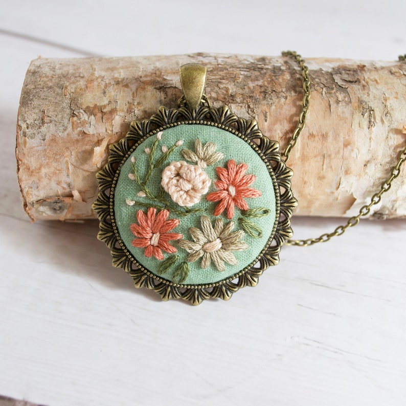 May include: A green fabric pendant with embroidered flowers in shades of pink, white, and green. The pendant is framed in a bronze-colored metal setting with a chain.