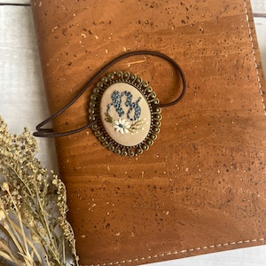 May include: A brown leather journal with a gold-toned metal charm featuring embroidered initials and floral details. The charm is attached to the journal with a brown elastic band.