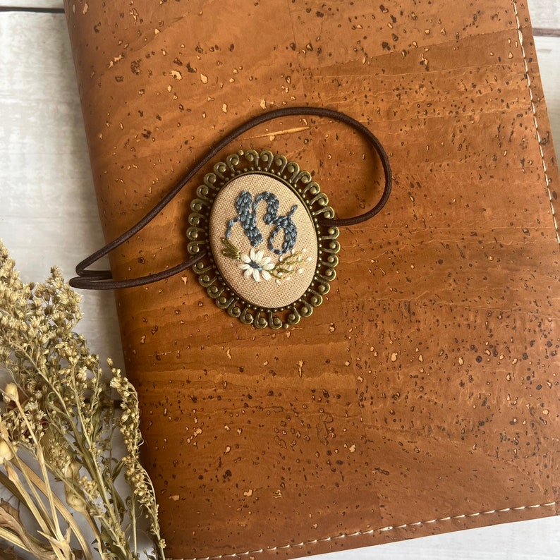 May include: A brown leather journal with a gold-toned metal charm featuring embroidered initials and floral details. The charm is attached to the journal with a brown elastic band.